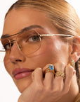 A woman with light skin and blonde hair wears gold-rimmed glasses and yellow gold rings, showcasing the Nijma M Fine Jewelry Yellow Gold Triple Band Cross Diamond Ring as she holds her hand near her face.