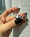 A hand with long, glossy black nails holds the Nijma M Fine Jewelry Marquise Diamond Eternity Ring in 18K white gold, with a customizable zigzag design, against a sheer white curtain in bright sunlight.