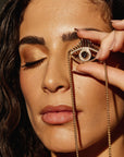 A woman with long dark hair holds the "Eye On You - Blue Topaz and Diamond Yellow Gold Evil Eye Necklace" from Nijma M Fine Jewelry in front of her closed eye. She has glowing, bronze-toned makeup and a neutral lip color, creating a striking, artistic effect. The dark background emphasizes her illuminated face.