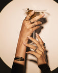 Two hands adorned with multiple rings and bracelets, including the "Asymmetrically Perfect - Marquise Diamond Open Front Yellow Gold Ring" from Nijma M Fine Jewelry, gracefully hold a martini glass with an olive garnish. The background features a large circular light, creating a dramatic and artistic shadow effect. The nails are elegantly manicured in a soft, neutral color.