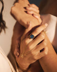 Close-up of intertwined hands, showcasing elegant jewelry. One hand features the stunning Blue Ice - Blue Topaz Triple Band Yellow Gold Ring by Nijma M Fine Jewelry and a delicate 14K yellow gold chain bracelet. The image captures a sense of intimacy and connection, highlighting the beautifully manicured nails and sophisticated accessories adorned with natural diamonds.