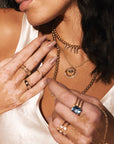 A close-up shot of a woman wearing multiple gold necklaces, including the "Guiding Light - Diamond Moon, Stars and Evil Eye Yellow Gold Necklace" by Nijma M Fine Jewelry and a rope chain necklace, along with rings like a blue gemstone ring and a pearl ring. She has light skin with visible patches of vitiligo, dark wavy hair, and is dressed in an ivory satin top. Her nails are painted nude.