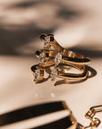 Close-up of the Asymmetrically Perfect - Marquise Diamond Open Front Yellow Gold Ring by Nijma M Fine Jewelry, showcasing its stylish gold design with three bands, each adorned with natural marquise-shaped diamonds that cast shadows on a light surface. Crafted from recycled 14K yellow gold, its open front design enhances its allure. In the foreground, a partially visible gold bracelet featuring black and white enamel sections is slightly out of focus.