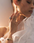 A woman wearing elegant jewelry poses with her eyes closed, head slightly tilted. Donning a white, ruffled sleeveless top with an open front design, she showcases several rings including the "Asymmetrically Perfect" Marquise Diamond Open Front Yellow Gold Ring by Nijma M Fine Jewelry. The soft lighting creates a warm and inviting atmosphere.