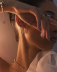 A person with hair tied back displays a graceful pose, partially covering their face with their hand. They wear elegant jewelry, including a gold bracelet, a necklace, and detailed yellow-gold earrings from Diamond Plaza - Square Hoop Diamond Earrings by Nijma M Fine Jewelry. The attire appears refined with a sheer, delicate fabric.