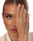 A blonde woman with natural makeup covers one eye and showcases the Nijma M Fine Jewelry White Gold Diamond Hand Chain, styled with a white top and gold hoop earring against a simple background.