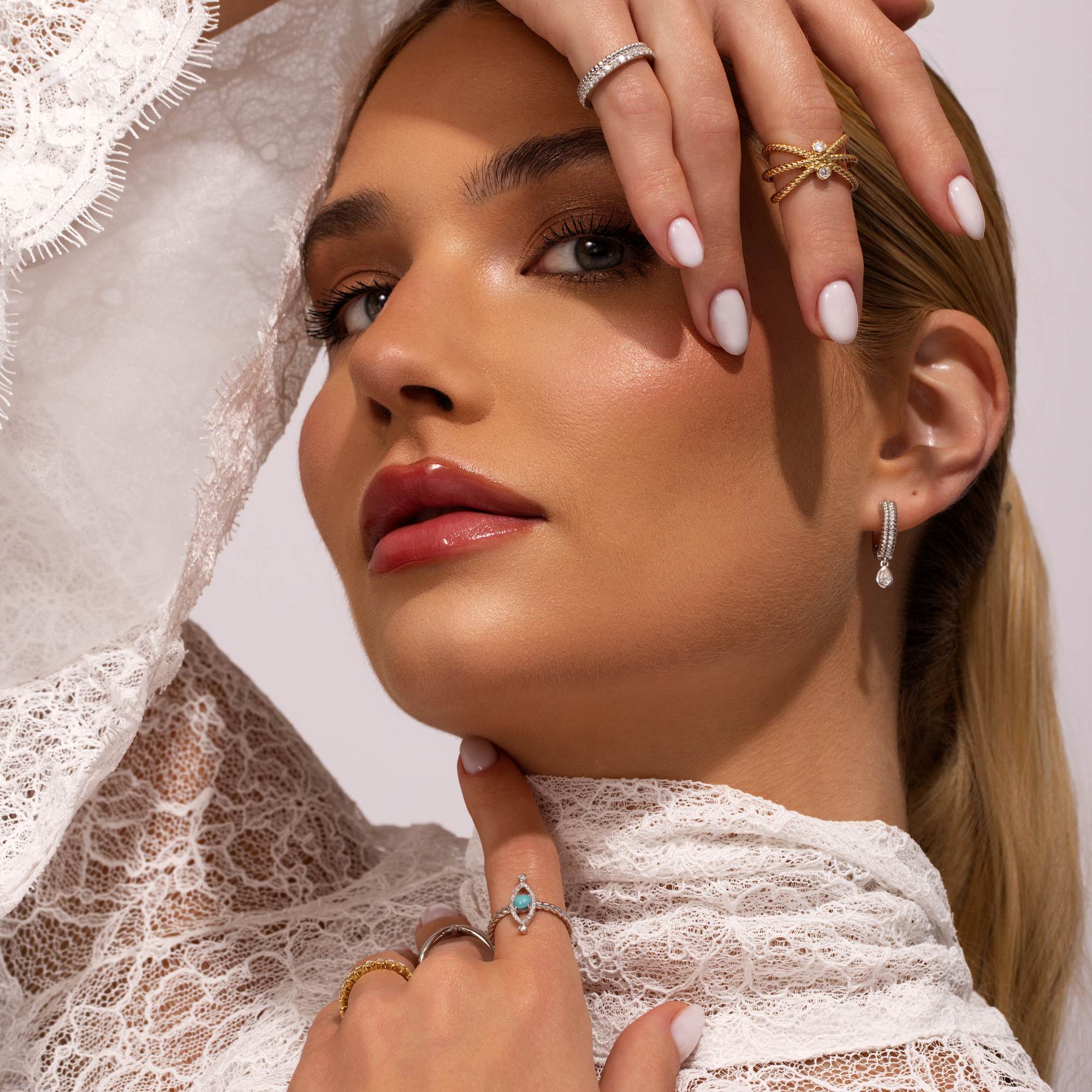 A woman in a white lace top poses with manicured white nails, stacked rings, and Nijma M Fine Jewelry's White Gold Fluted Hoop Earrings with Dangling Diamonds. Her polished makeup complements her confident gaze at the camera.