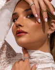 A woman in a white lace top poses with manicured white nails, stacked rings, and Nijma M Fine Jewelry's White Gold Fluted Hoop Earrings with Dangling Diamonds. Her polished makeup complements her confident gaze at the camera.