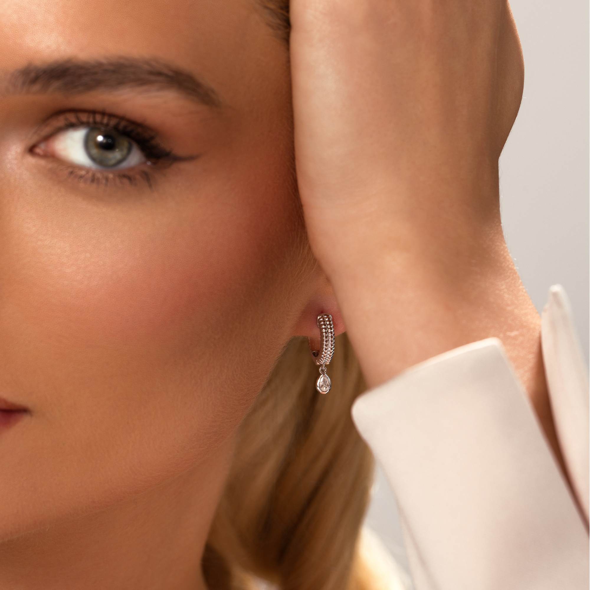 Close-up of a woman with blue eyes and a white blouse wearing Nijma M Fine Jewelry’s White Gold Fluted Hoop Earrings with Dangling Diamonds. She touches her hair, highlighting the sparkling diamond hoops.