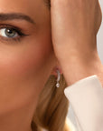 Close-up of a woman with blue eyes and a white blouse wearing Nijma M Fine Jewelry’s White Gold Fluted Hoop Earrings with Dangling Diamonds. She touches her hair, highlighting the sparkling diamond hoops.