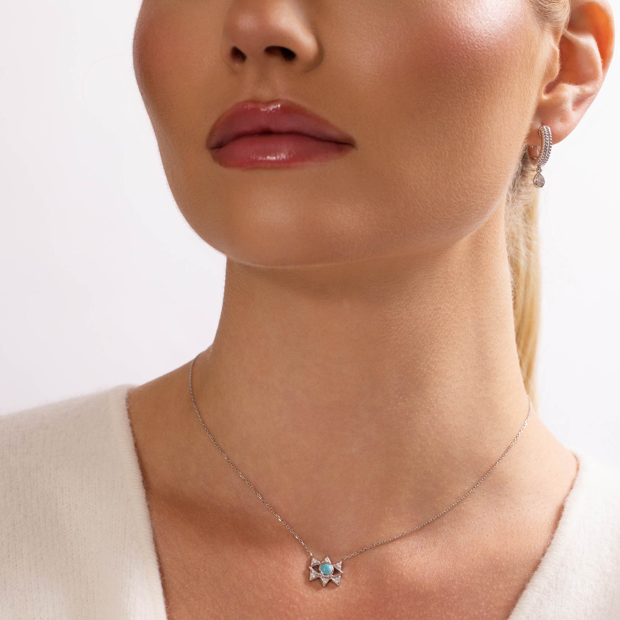 A woman with glossy lips and pulled-back hair is shown from the shoulders up, modeling the Nijma M Fine Jewelry Small White Gold Evil Eye Necklace and a small earring. She wears a white top.