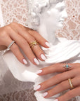 A woman's manicured hands, adorned with Nijma M Fine Jewelry's Yellow Gold Triple Band Cross Diamond Ring and other elegant rings in gold, silver, diamonds, and turquoise, rest on a white marble bust against lace.