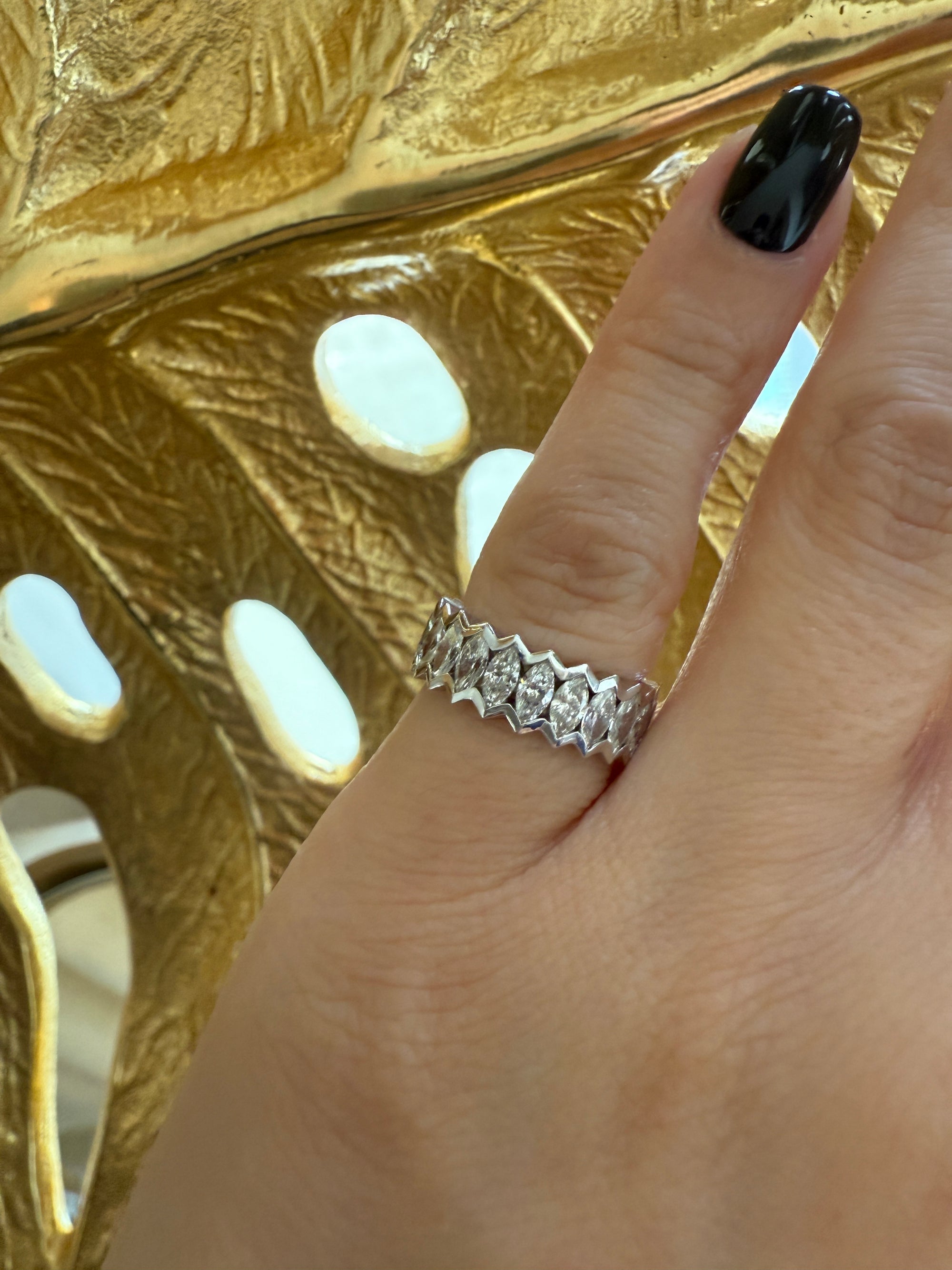 A hand with black nail polish wears Nijma M Fine Jewelry’s Marquise Diamond Eternity Ring, featuring marquise-shaped diamonds, set against a gold background with cut-out designs.