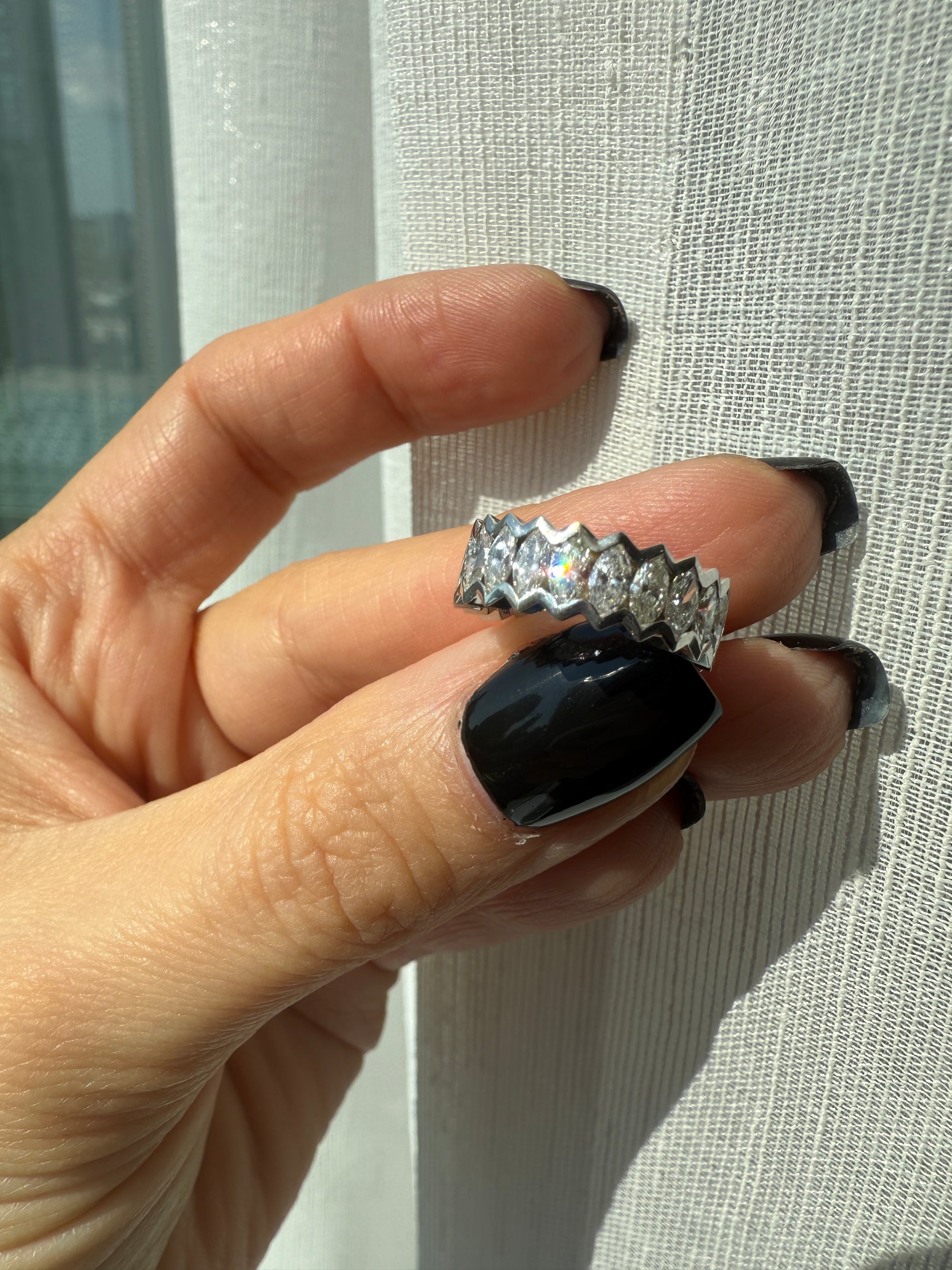 A hand with long, glossy black nails holds the Nijma M Fine Jewelry Marquise Diamond Eternity Ring in 18K white gold, with a customizable zigzag design, against a sheer white curtain in bright sunlight.