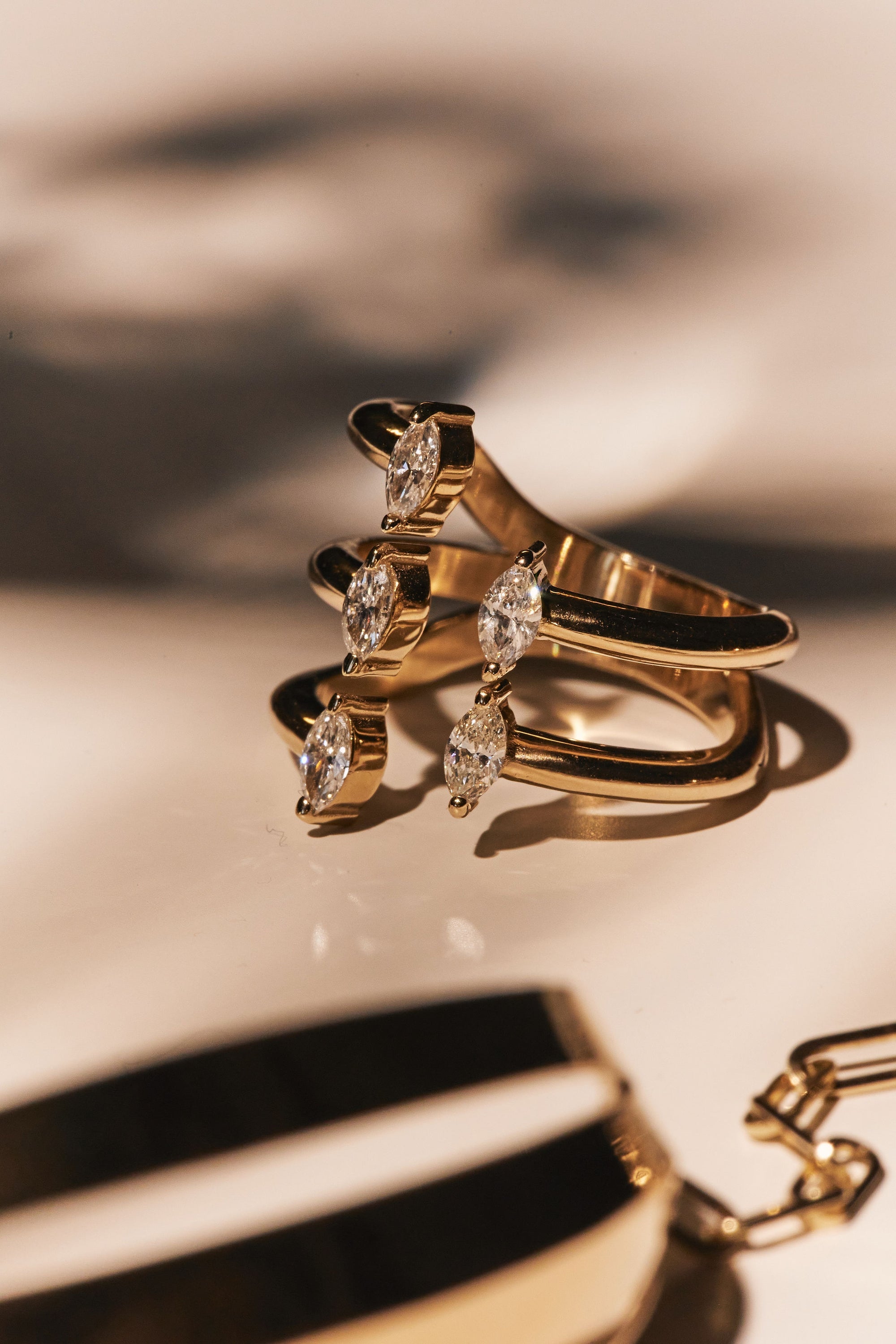 Close-up of the Asymmetrically Perfect - Marquise Diamond Open Front Yellow Gold Ring by Nijma M Fine Jewelry, showcasing its stylish gold design with three bands, each adorned with natural marquise-shaped diamonds that cast shadows on a light surface. Crafted from recycled 14K yellow gold, its open front design enhances its allure. In the foreground, a partially visible gold bracelet featuring black and white enamel sections is slightly out of focus.