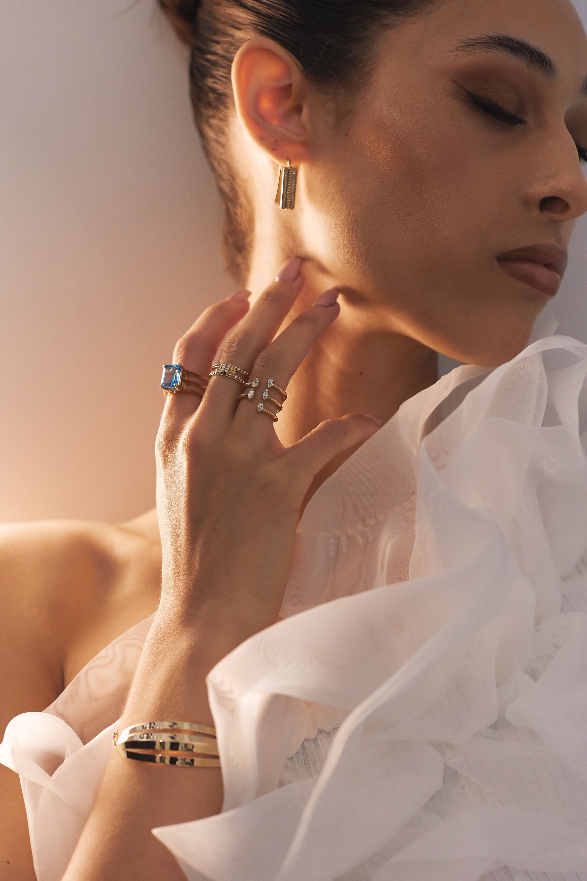 A woman with her eyes closed wears gold earrings, multiple rings, including the "Golden Baguette - Yellow Gold Baguette Diamond Ring" by Nijma M Fine Jewelry, and an elegant gold bracelet. She is dressed in a white ruffled top and touches her neck with her right hand. Golden light illuminates her face and hand.