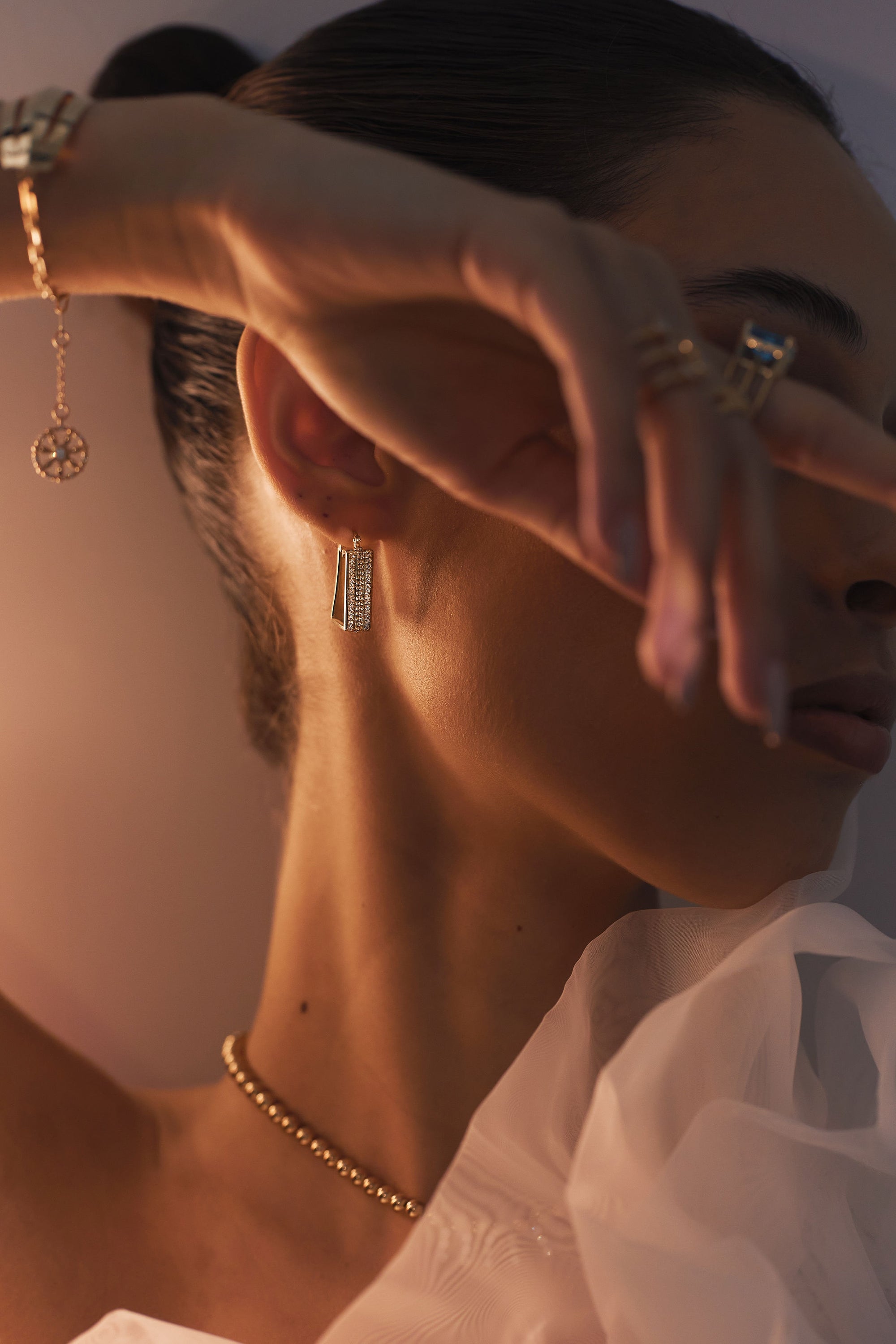 A person with hair tied back displays a graceful pose, partially covering their face with their hand. They wear elegant jewelry, including a gold bracelet, a necklace, and detailed yellow-gold earrings from Diamond Plaza - Square Hoop Diamond Earrings by Nijma M Fine Jewelry. The attire appears refined with a sheer, delicate fabric.