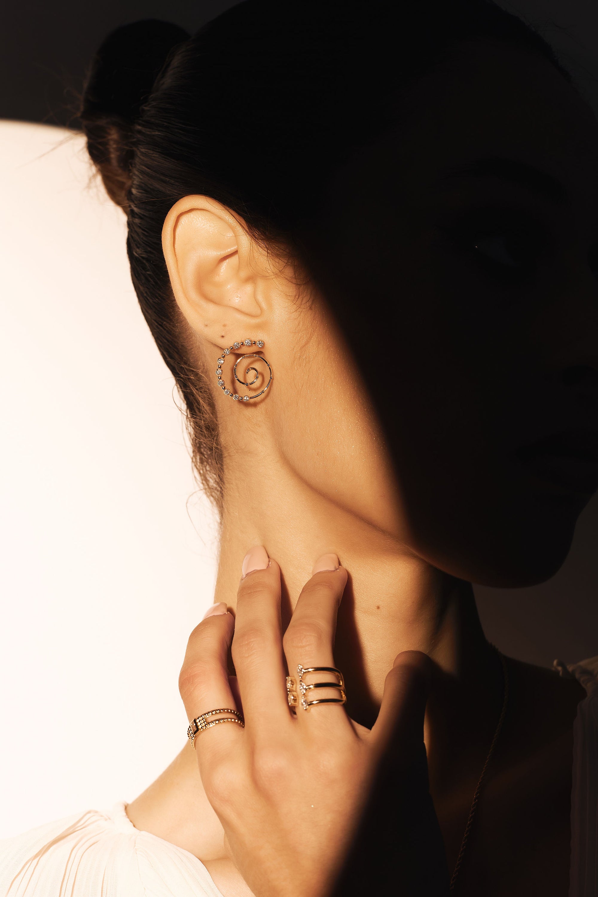 A close-up photo captures a person in partial shadow wearing Spiralize - Diamond And Yellow Gold Spiral Earrings from Nijma M Fine Jewelry, along with multiple rings adorned with diamond beads. The person's hair is tied up, and they are dressed in a light-colored top, gently touching the side of their neck. The dramatic lighting emphasizes the handmade craftsmanship of the jewelry.