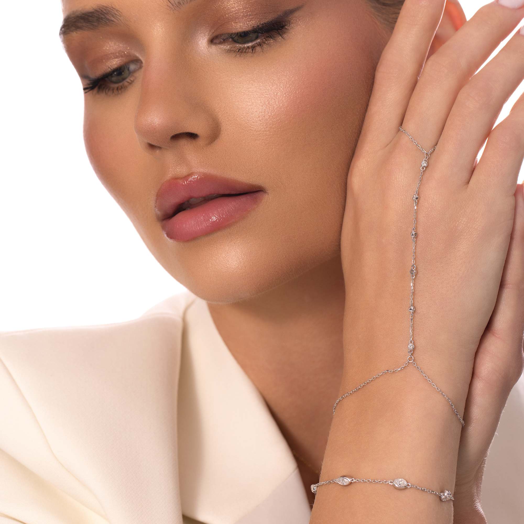 A woman in a cream blazer poses with her hand near her face, highlighting the White Gold Diamond Station Bracelet by Nijma M Fine Jewelry. Her soft, natural makeup features glossy lips and subtle eye shadow.
