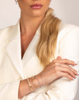 A woman with long blonde hair in a ponytail, wearing gold and silver bracelets—including the Nijma M Fine Jewelry White Gold Diamond Station Bracelet—poses with arms crossed against a plain background.