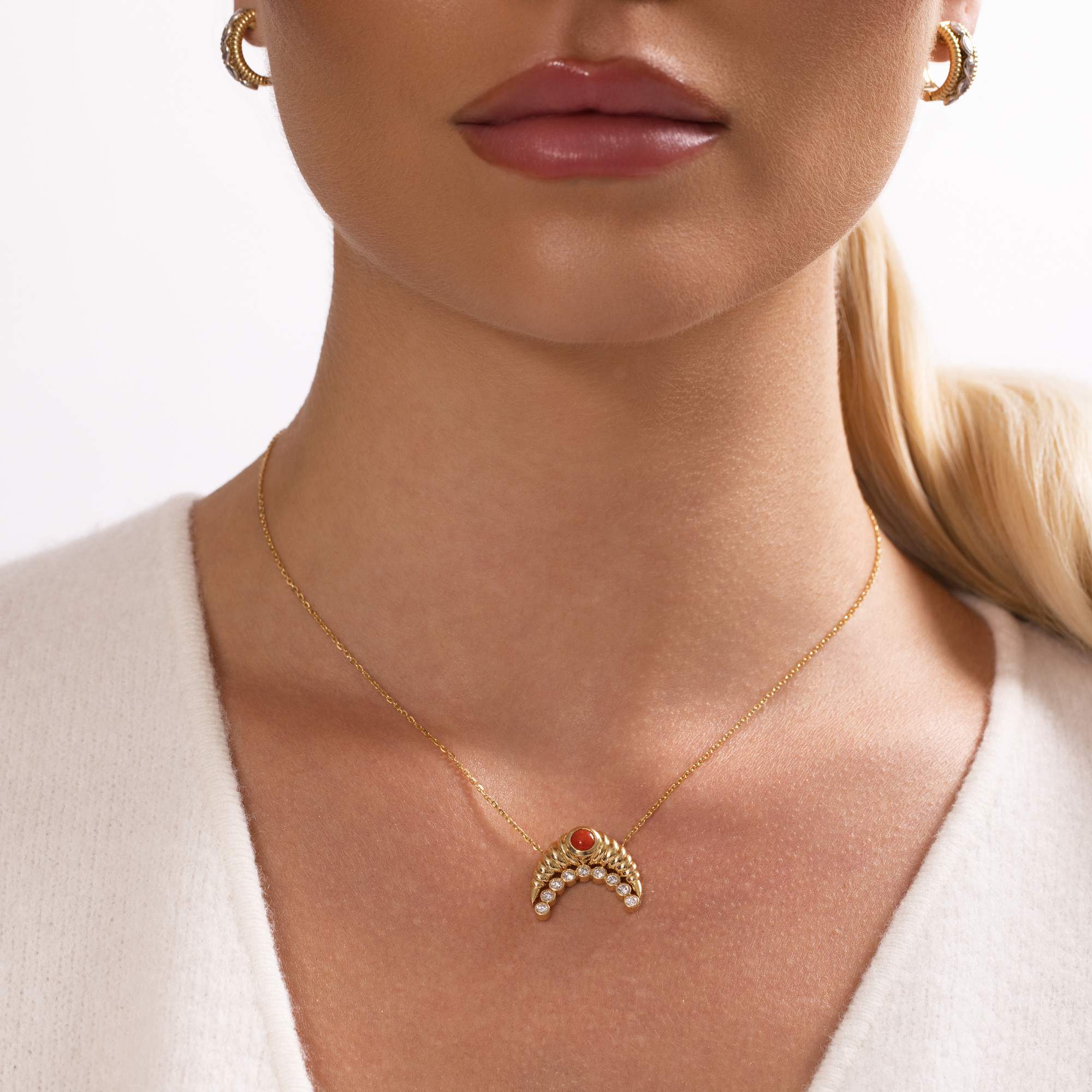 A woman with blond hair in a low ponytail wears Nijma M Fine Jewelry’s Diamond and Coral Gold Moon Necklace and matching gold hoop earrings. She has light skin, glossy lips, and a white top.