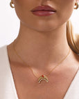 A woman with blond hair in a low ponytail wears Nijma M Fine Jewelry’s Diamond and Coral Gold Moon Necklace and matching gold hoop earrings. She has light skin, glossy lips, and a white top.