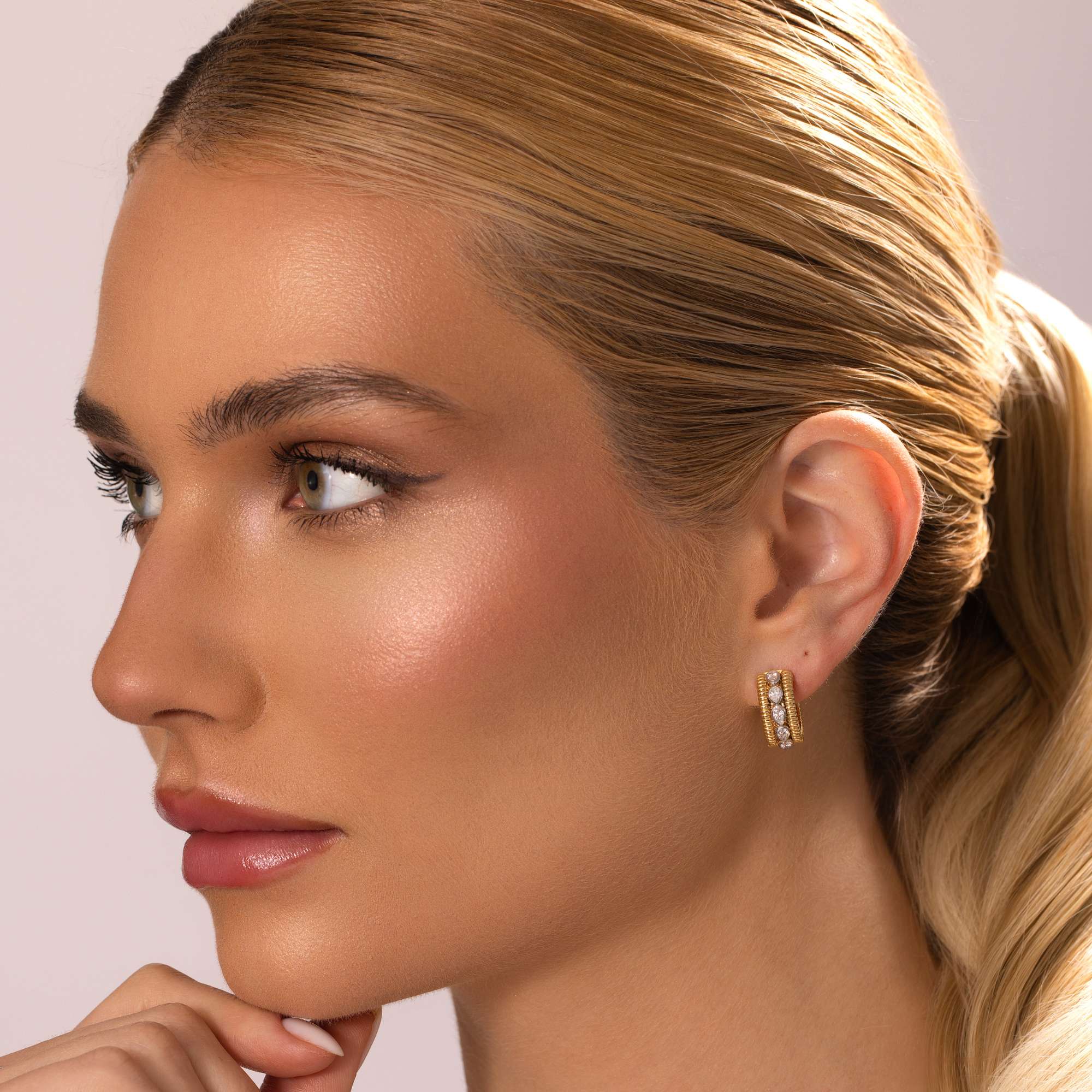 A woman with blonde hair in a sleek ponytail wears Nijma M Fine Jewelry's Small Diamond Hoop Earrings. Her smooth, glowing skin and natural makeup enhance her look as she gazes left against a light background.