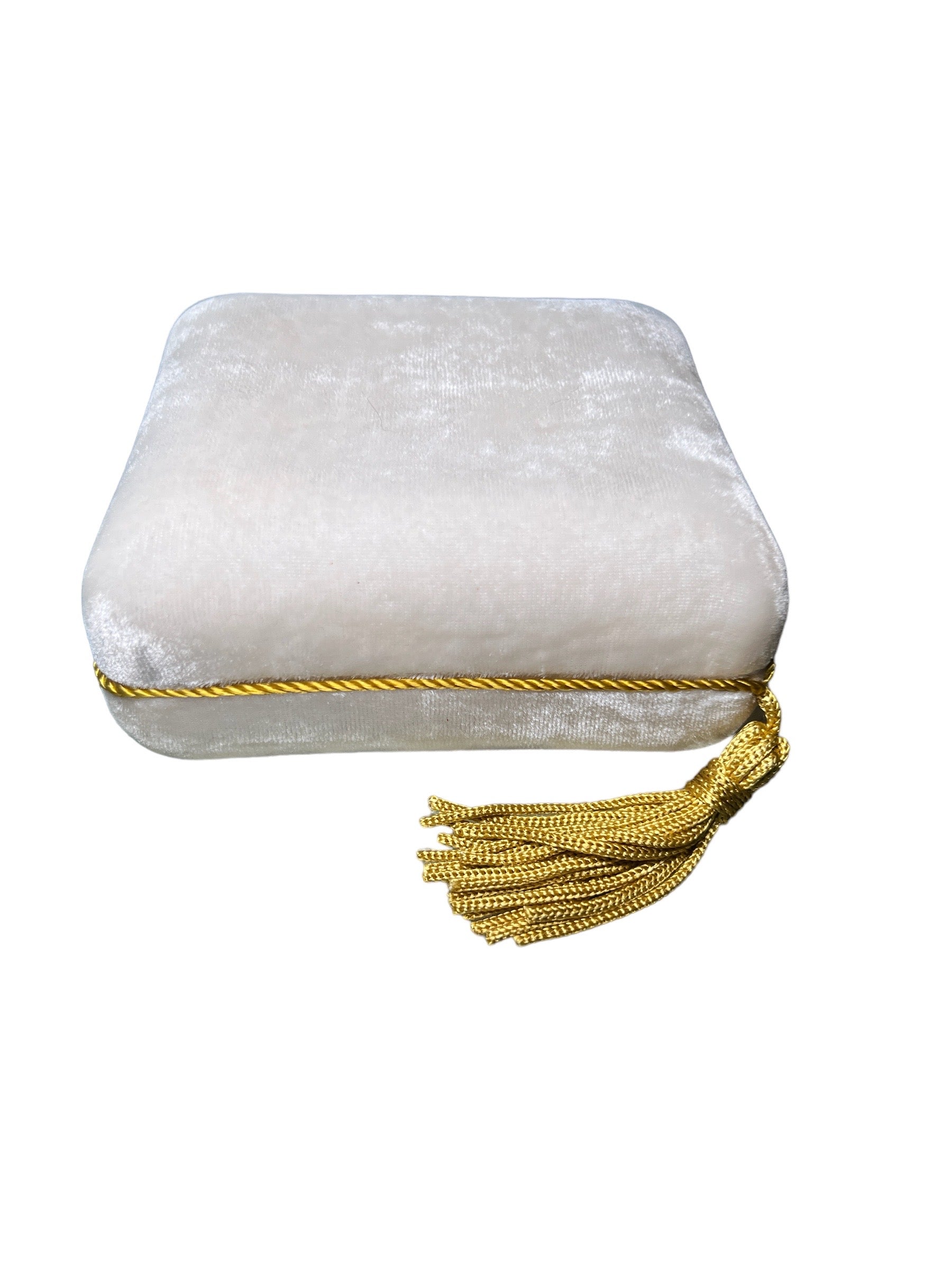 The Nijma M Fine Jewelry Pochette Case is a rectangular, white crushed velvet case with a rounded top. It is embellished with a gold braided trim around its edge and features elegant gold tassel detailing hanging from one corner. The background is plain white.