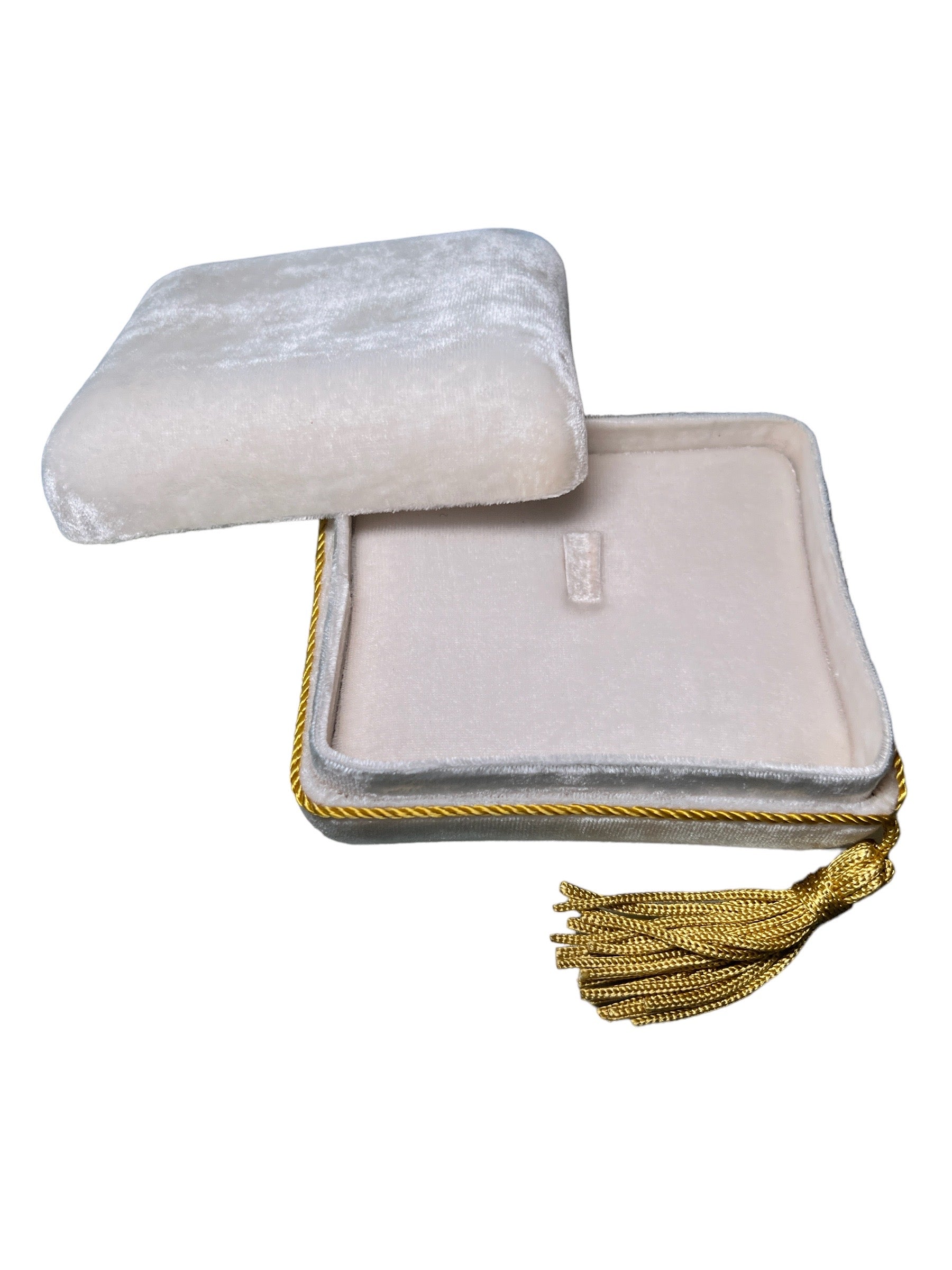 The Jewelry Pochette Case by Nijma M Fine Jewelry is open, showcasing its soft, plush velvet interior. The square box's lid is slightly askew, while a gold tassel detailing elegantly hangs from one corner. Both the exterior and interior are crafted from a light cream-colored velvet fabric.
